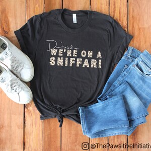 May include: Black t-shirt with a leopard print graphic that says "Don't mind us we're on a sniffari". The shirt is laid out on a wooden surface with a pair of white sneakers and blue jeans.