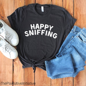 Può includere: Maglietta nera con testo bianco che dice "HAPPY SNIFFING" su uno sfondo di legno con jeans blu e scarpe da ginnastica bianche.