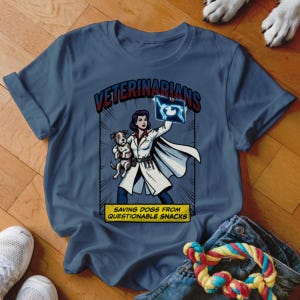 Puede incluir: Una camiseta azul con una ilustración estilo cómic de un veterinario sosteniendo una radiografía y un perro pequeño. El texto "VETERINARIANS" está encima de la imagen y "SAVING DOGS FROM QUESTIONABLE SNACKS" debajo.
