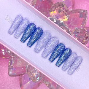 May include: A set of ten false nails with a gradient of light blue and purple glitter. The nails are arranged in a box.