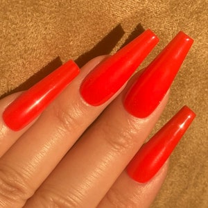 May include: Close-up of a hand with long, bright red, coffin-shaped acrylic nails. The nails are glossy and reflect light, creating highlights. The background is a textured, tan surface.
