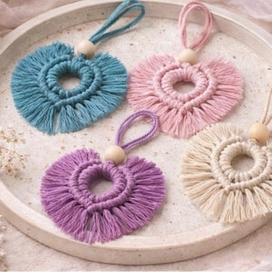 Może przedstawiać: Cztery makramowe ozdoby w kształcie serca w odcieniach turkusu, różu, fioletu i kremu, każda z pętelką do zawieszenia i drewnianą koralikiem. Serca ułożone są na okrągłej, kremowej tacy.