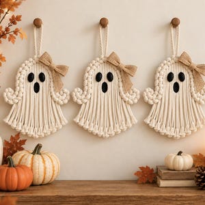 May include: Three off-white macrame ghost wall hangings, each featuring a tan burlap bow, two black oval eyes, and a black oval mouth. The ghosts are suspended by twisted rope loops from wooden wall pegs, creating a uniform set of decorations.