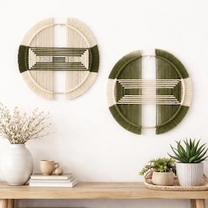 Puede incluir: Dos colgantes de pared circulares con un diseño tejido. Cada uno presenta un anillo exterior color crema, una sección verde oscuro y un centro geométrico crema y verde. Los colgantes están montados en una pared blanca sobre una mesa de madera.