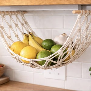 May include: A macrame fruit hammock, suspended from a wooden rod, filled with yellow lemons, green limes, and bananas. A white garlic bulb is also visible. The hammock hangs in front of a white tiled wall.