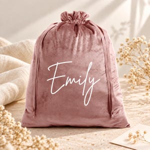 Puede incluir: Una bolsa de terciopelo color rosa empolvado con cierre de cordón que presenta el nombre Emily escrito en letra cursiva blanca. La bolsa tiene un cierre fruncido en la parte superior con cintas de terciopelo a juego, ofreciendo una textura suave.