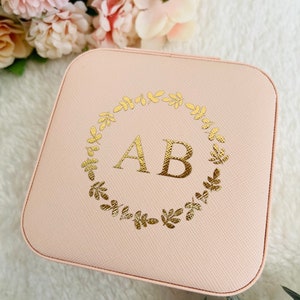 以下が含まれることがあります： 薄いピンク色の正方形のジュエリーボックス。金箔で葉の輪と「AB」のイニシャルがデザインされています。