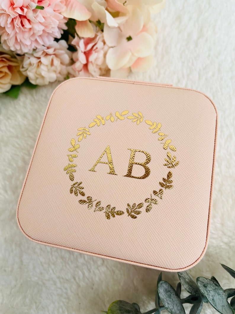 以下が含まれることがあります： 薄いピンク色の正方形のジュエリーボックス。金箔で葉の輪と「AB」のイニシャルがデザインされています。