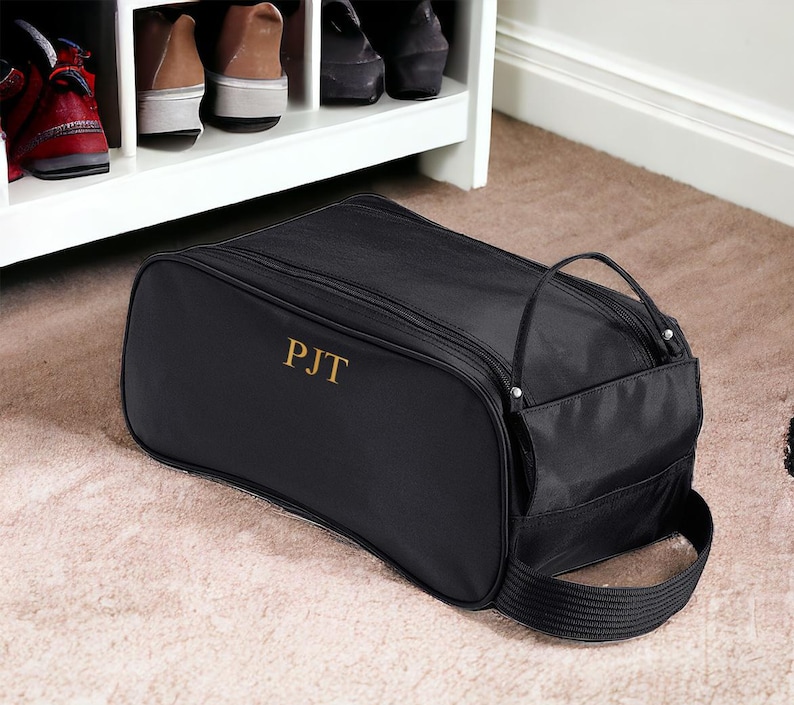 May include: A black rectangular shoe bag with a zipped closure and a side handle. The bag features the gold embroidered letters PJT on the front panel. The exterior is made of a smooth fabric, and the design includes a structured shape for storage.