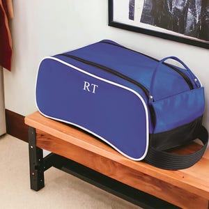May include: A royal blue duffel bag with white piping trim and black accents. The bag features a zippered main compartment and a side pocket, with the white letters RT monogrammed on the front side panel of the bag's exterior fabric.