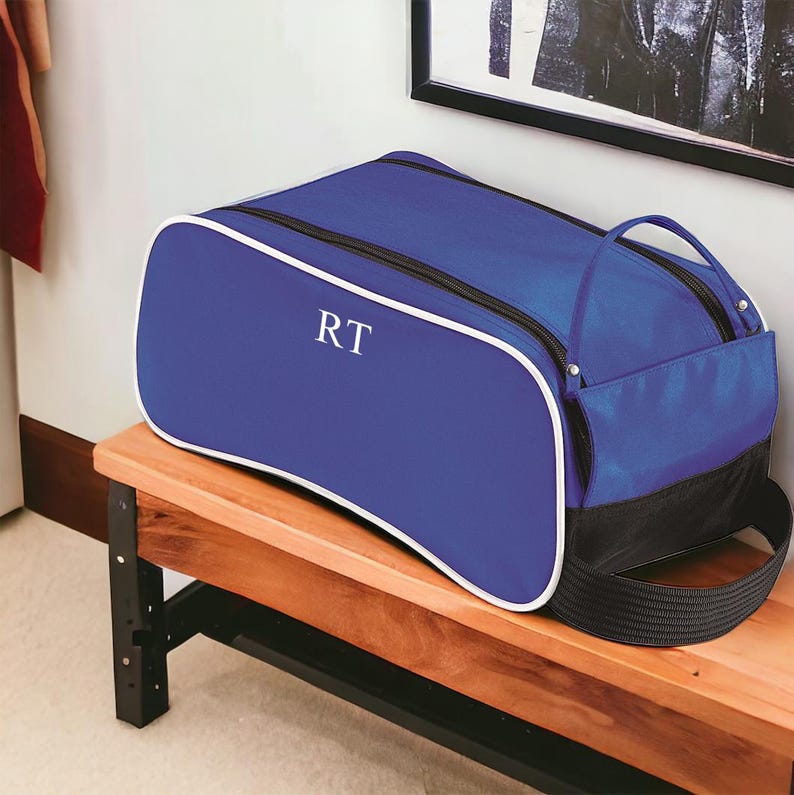 May include: A royal blue duffel bag with white piping trim and black accents. The bag features a zippered main compartment and a side pocket, with the white letters RT monogrammed on the front side panel of the bag's exterior fabric.