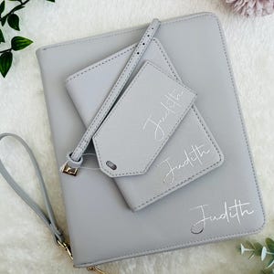 May include: A light gray personalized clutch, wallet, and luggage tag set. The clutch has a wrist strap and gold zipper. The wallet and luggage tag are stacked on top. The name "Judith" is written in silver script on each item.