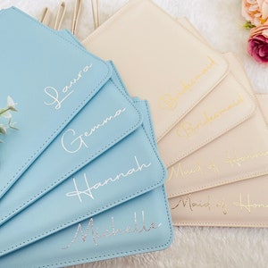May include: Five light blue and beige clutch bags with different gold and silver lettering. The bags are labeled "Laura", "Gemma", "Hannah", "Michelle", "Bridesmaid", "Maid of Honor", and "Maid of Honor".