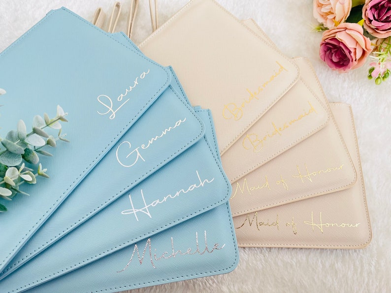 May include: Five light blue and beige clutch bags with different gold and silver lettering. The bags are labeled "Laura", "Gemma", "Hannah", "Michelle", "Bridesmaid", "Maid of Honor", and "Maid of Honor".