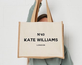May include: A rectangular tote bag made of off-white canvas with tan jute side panels and matching handles. The front features black printed text that reads N°40, KATE WILLIAMS, and LONDON in a clean, sans-serif font centered on the bag surface.