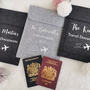 May include: Three gray felt travel document holders with white text and a small airplane graphic. The holders are labeled "The Martin's Travel Documents", "The Bakewell's Familymoon 2022", and "The King's Travel Documents". Two passports are visible in the foreground.