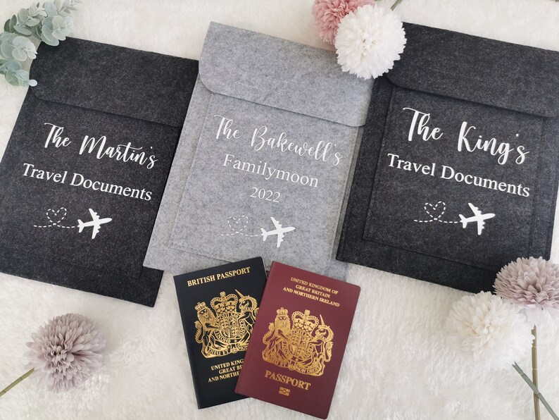 May include: Three gray felt travel document holders with white text and a small airplane graphic. The holders are labeled "The Martin's Travel Documents", "The Bakewell's Familymoon 2022", and "The King's Travel Documents". Two passports are visible in the foreground.