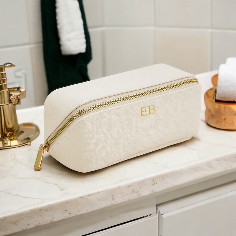 May include: Cream-colored rectangular cosmetic bag with a gold zipper and pull. The bag has the gold initials "EB" on the front. The bag is sitting on a white marble countertop.