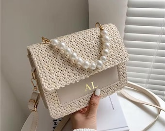 Peut inclure: Un sac à bandoulière en paille tressée beige doté d'un rabat avec une poignée décorative en perles blanches. Le panneau avant comprend un insert beige lisse avec les lettres dorées AL imprimées au centre. Il possède une bandoulière assortie.