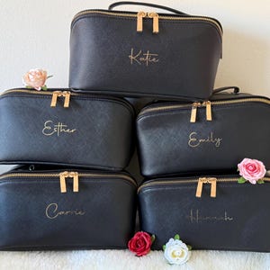 Pu&ograve; includere: Cinque trousse per cosmetici nere con cerniere e manici dorati sono impilate. Ogni borsa ha un nome in oro: Katie, Esther, Emily, Carrie e Hannah. Fiori rosa e rossi sono posti intorno alle borse.