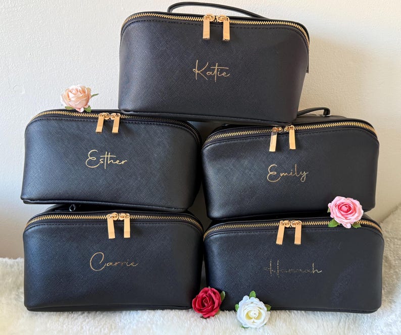 Pu&ograve; includere: Cinque trousse per cosmetici nere con cerniere e manici dorati sono impilate. Ogni borsa ha un nome in oro: Katie, Esther, Emily, Carrie e Hannah. Fiori rosa e rossi sono posti intorno alle borse.