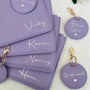 May include: Five purple zippered pouches with gold zippers and matching round keychains. The pouches are personalized with the names "Vicky", "Karen", "Nanny", "Hannah", and "Bridesmaid". The keychains are also personalized with the names "Bridesmaid", "Flower Girl", and "Bridesmaid".