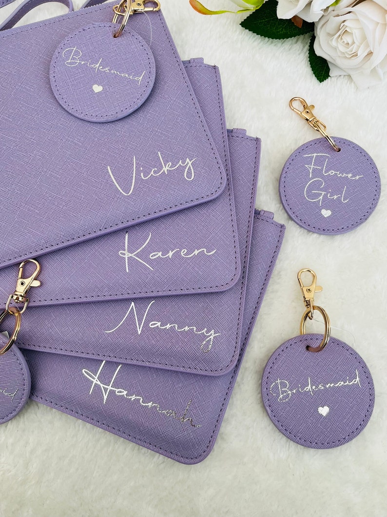 May include: Five purple zippered pouches with gold zippers and matching round keychains. The pouches are personalized with the names "Vicky", "Karen", "Nanny", "Hannah", and "Bridesmaid". The keychains are also personalized with the names "Bridesmaid", "Flower Girl", and "Bridesmaid".