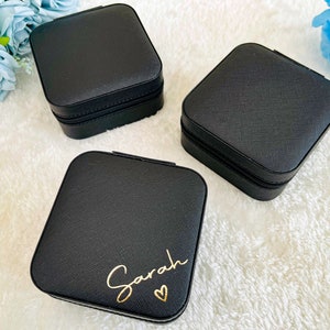 K&ouml;nnte beinhalten: Drei schwarze quadratische Schmuckk&auml;stchen mit Rei&szlig;verschluss. Eine Box tr&auml;gt den Namen "Sarah" in goldenen Lettern mit einem kleinen Herz.