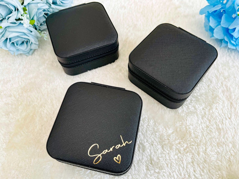 K&ouml;nnte beinhalten: Drei schwarze quadratische Schmuckk&auml;stchen mit Rei&szlig;verschluss. Eine Box tr&auml;gt den Namen "Sarah" in goldenen Lettern mit einem kleinen Herz.