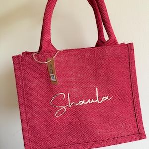 May include: A red burlap tote bag with a matching handle. The bag features the name "Shaula" in a gold glitter script. A small tag hangs from the handle.
