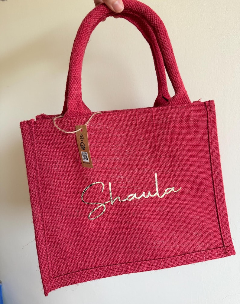 May include: A red burlap tote bag with a matching handle. The bag features the name "Shaula" in a gold glitter script. A small tag hangs from the handle.