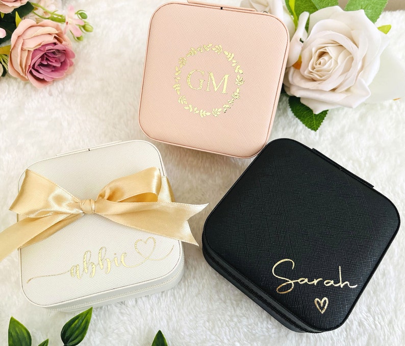 K&ouml;nnte beinhalten: Drei verschiedene Schmuckk&auml;stchen mit personalisierten Gravuren. Die erste Schachtel ist rosa mit einem goldenen Blumengirlanden und den Initialen "GM". Die zweite Schachtel ist wei&szlig; mit einer goldenen Schleife und dem Namen "abbie" in Kursivschrift. Die dritte Schachtel ist schwarz mit dem Namen "Sarah" in Kursivschrift.