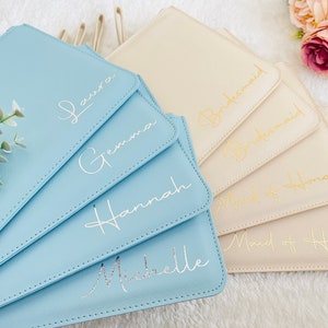 May include: A collection of rectangular pouches in light blue and cream colors, featuring stitched edges and gold or silver cursive lettering. The blue pouches are personalized with the names Laura, Gemma, Hannah, and Michelle, while the cream pouches are labeled with the titles Bridesmaid and Maid of Honour.