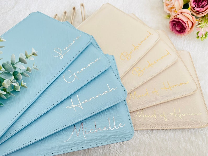 May include: A collection of rectangular pouches in light blue and cream colors, featuring stitched edges and gold or silver cursive lettering. The blue pouches are personalized with the names Laura, Gemma, Hannah, and Michelle, while the cream pouches are labeled with the titles Bridesmaid and Maid of Honour.