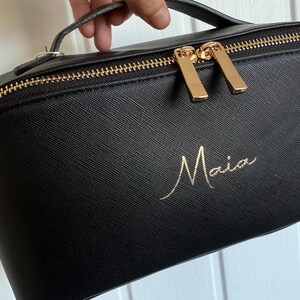 May include: Black makeup bag with a gold zipper and handle. The bag has the name "Maia" in gold script on the front. The zipper pulls are gold.