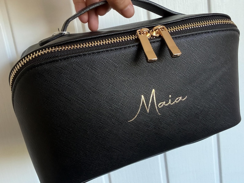May include: Black makeup bag with a gold zipper and handle. The bag has the name "Maia" in gold script on the front. The zipper pulls are gold.