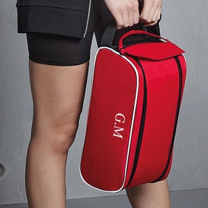May include: A red rectangular zipped bag with white piping trim and a black handle. The bag features the white embroidered letters G.M on the front panel. The design includes a top flap closure and black zip accents along the sides.