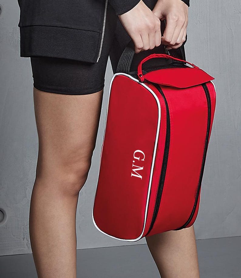 May include: A red rectangular zipped bag with white piping trim and a black handle. The bag features the white embroidered letters G.M on the front panel. The design includes a top flap closure and black zip accents along the sides.