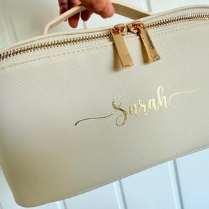 May include: Cream-colored cosmetic bag with a gold zipper and handle. The bag features the name "Sarah" in gold script. The zipper pulls are gold, and the bag has a textured surface.
