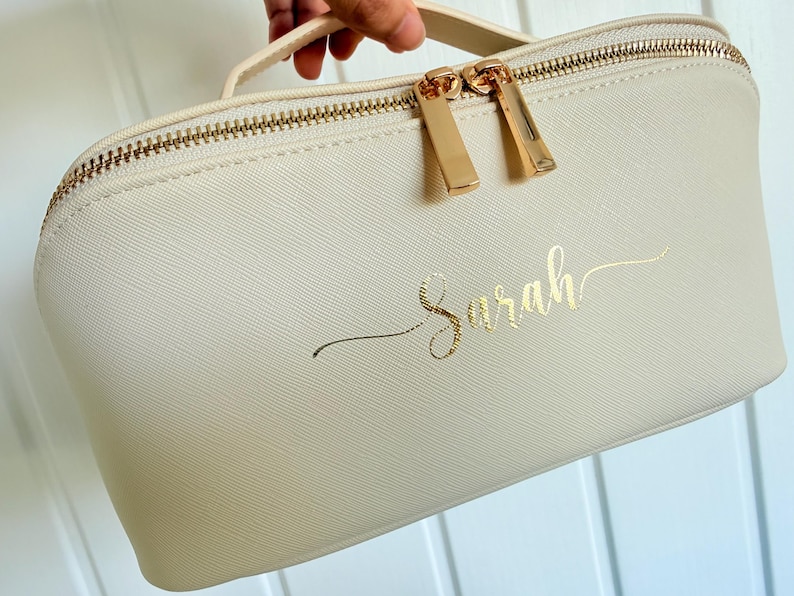 May include: Cream-colored cosmetic bag with a gold zipper and handle. The bag features the name "Sarah" in gold script. The zipper pulls are gold, and the bag has a textured surface.