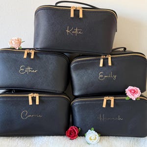 May include: Black cosmetic bags with gold zippers and handles are stacked. Each bag has a name in gold script: Katie, Esther, Emily, Carrie, and Hannah. Pink and white flowers are placed around the bags.