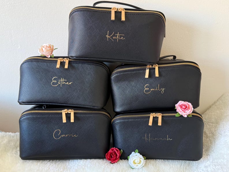May include: Black cosmetic bags with gold zippers and handles are stacked. Each bag has a name in gold script: Katie, Esther, Emily, Carrie, and Hannah. Pink and white flowers are placed around the bags.