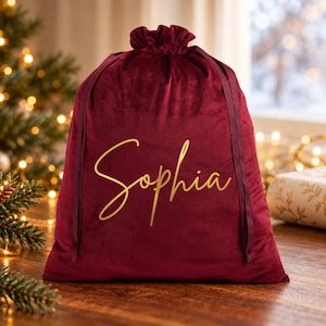 May include: A deep red velvet drawstring gift bag featuring the name Sophia written in gold cursive script. The bag has a gathered top closure with matching red ribbon ties and a soft, plush texture throughout the fabric surface.