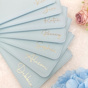 May include: A stack of light blue zippered pouches with gold lettering. The pouches are personalized with the names Lexie, Jess, Katie, Becky, Sophie, Alison, and Debbie.