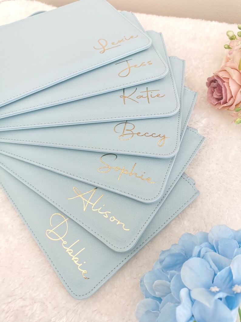 May include: A stack of light blue zippered pouches with gold lettering. The pouches are personalized with the names Lexie, Jess, Katie, Becky, Sophie, Alison, and Debbie.