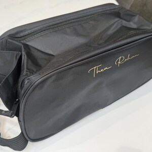 May include: A black rectangular travel bag with a zipped closure and a black fabric shoulder strap. The side of the bag features the name Thea Rohan embroidered in a gold cursive script font. The bag is constructed from a smooth, durable material.