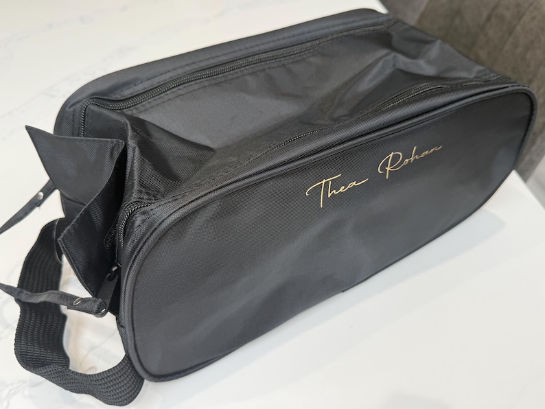 May include: A black rectangular travel bag with a zipped closure and a black fabric shoulder strap. The side of the bag features the name Thea Rohan embroidered in a gold cursive script font. The bag is constructed from a smooth, durable material.