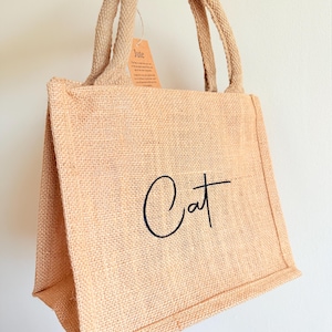 May include: A beige jute tote bag with the word "Cat" in black script. The bag has two handles and a small tag that reads "Jute". The bag is made of a woven material.