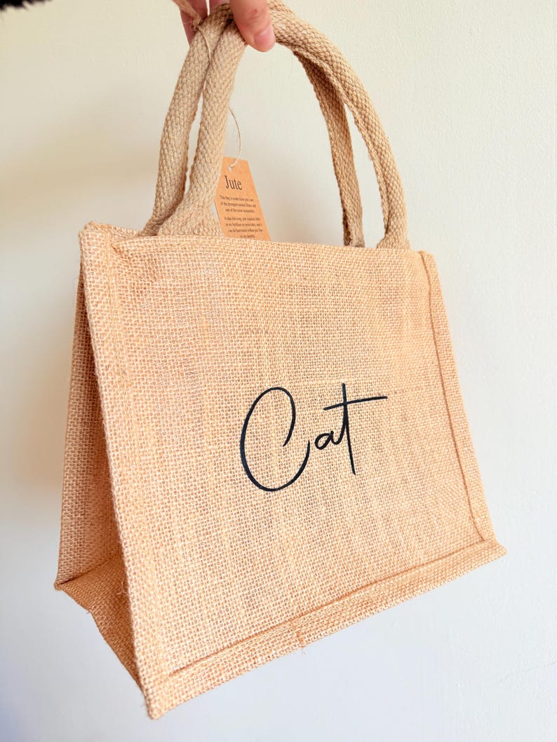 May include: A beige jute tote bag with the word "Cat" in black script. The bag has two handles and a small tag that reads "Jute". The bag is made of a woven material.