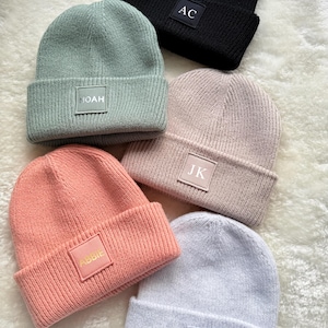 Pode incluir: Uma coleção de cinco gorros de malha em várias cores: preto, verde sálvia, rosa claro, bege e cinza claro. Cada gorro apresenta um patch retangular com texto personalizado: "AC", "NOAH", "ABIE", "JK" e "F.B".